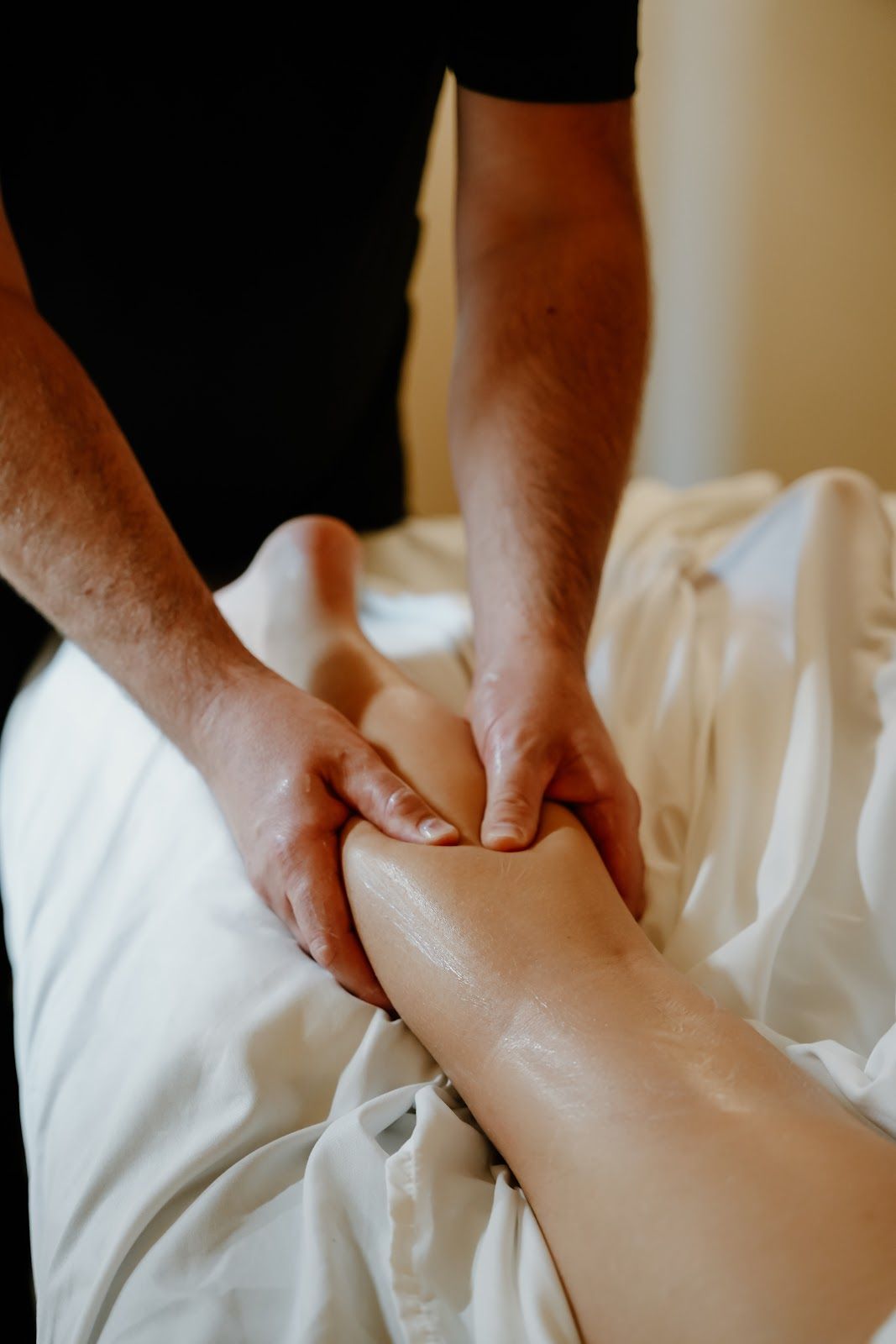 Person massaging a leg on a massage table, using both hands. The leg is oiled and on white sheets.