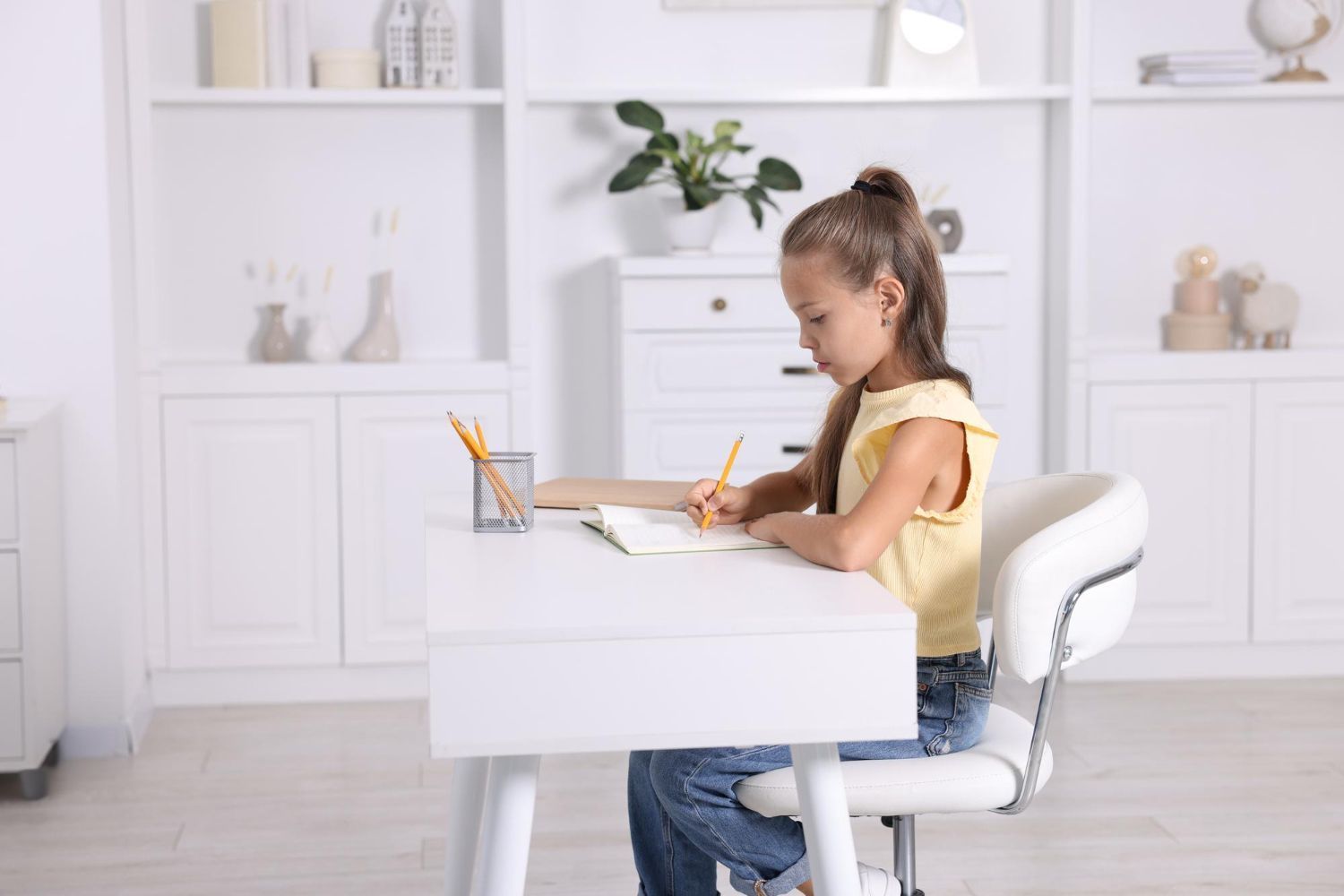 Girl writing at a white desk in a bright room. She wears a yellow top and jeans.