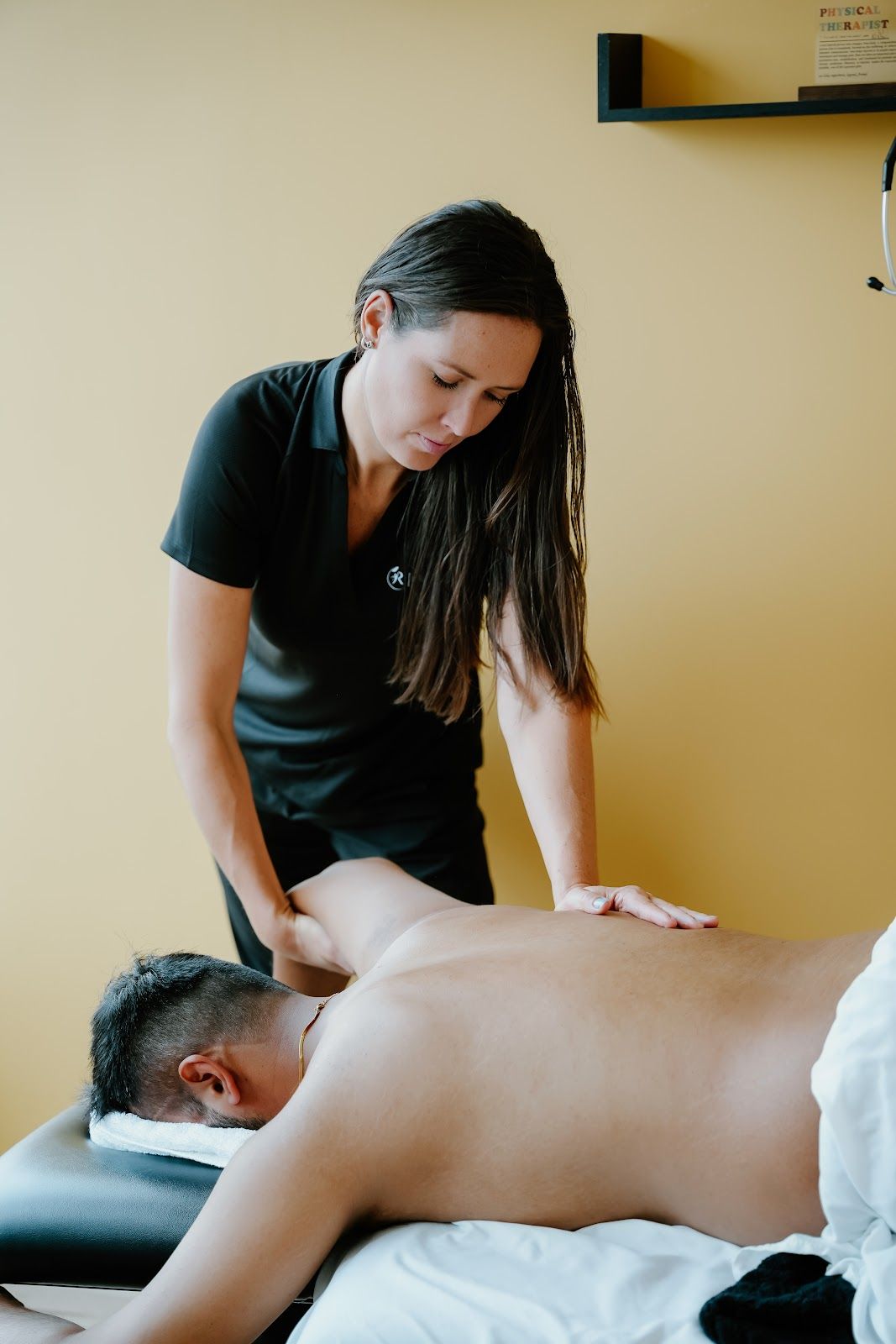 Massage therapist working on a person's back; tan walls, black top.