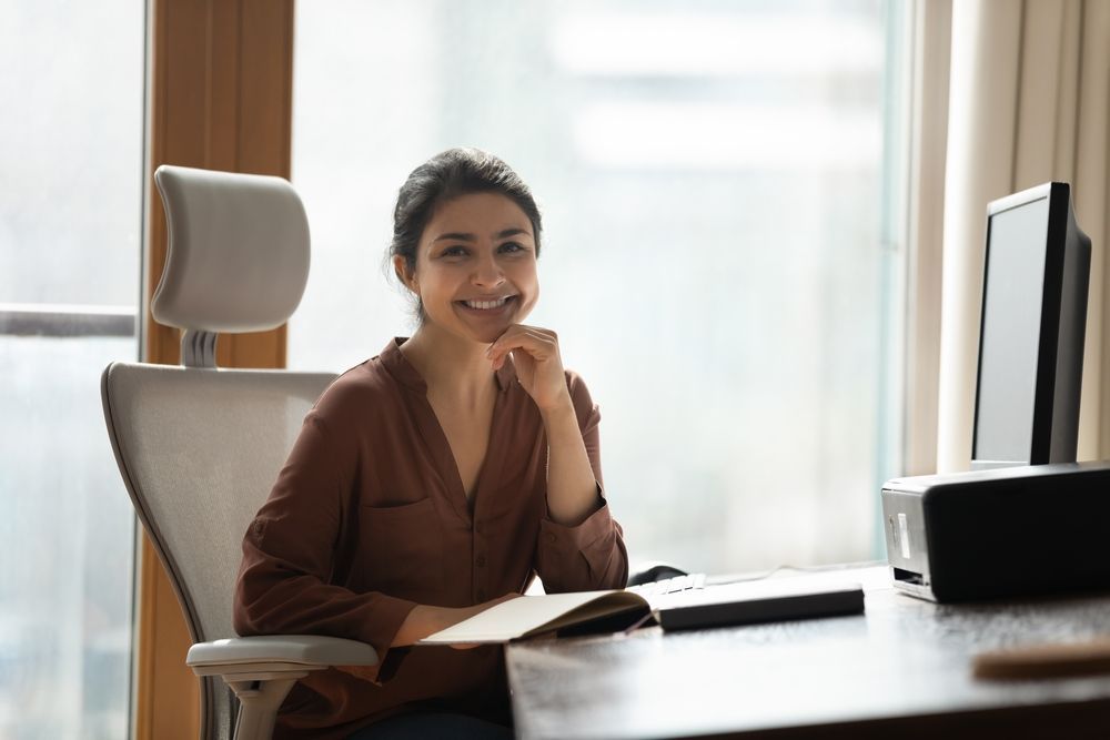 A smiling professional sits at a desk with an open notebook, facing a computer monitor in a bright, modern office.
