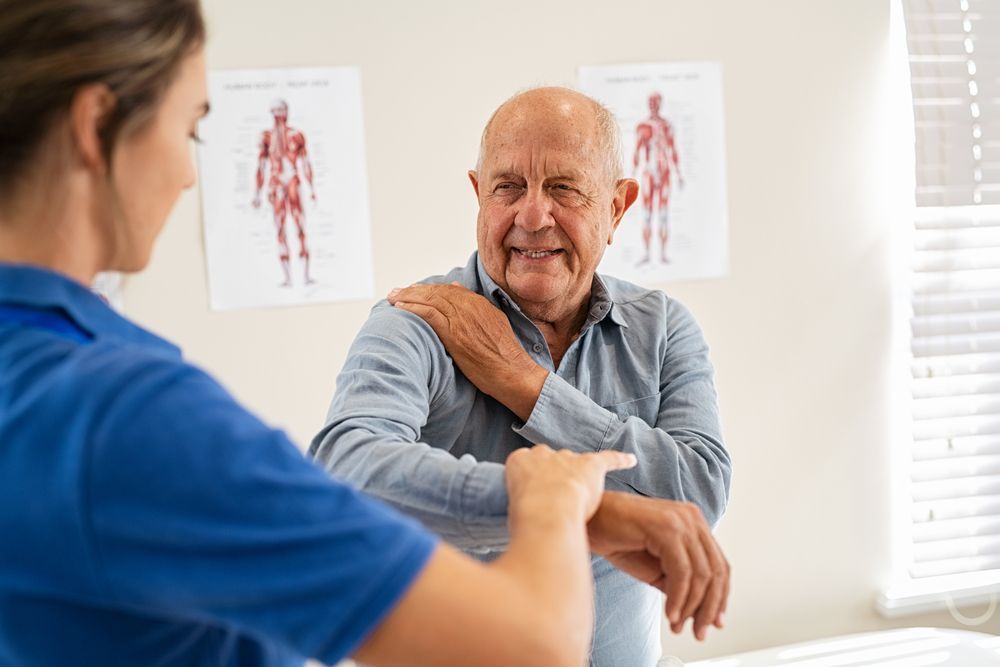 A professional in a blue shirt performs a physical examination on a person who is holding their shoulder in a clinic.