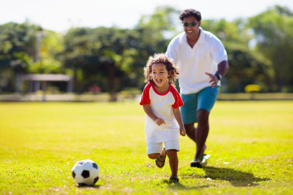 A child and adult run together across a grassy field chasing a soccer ball.