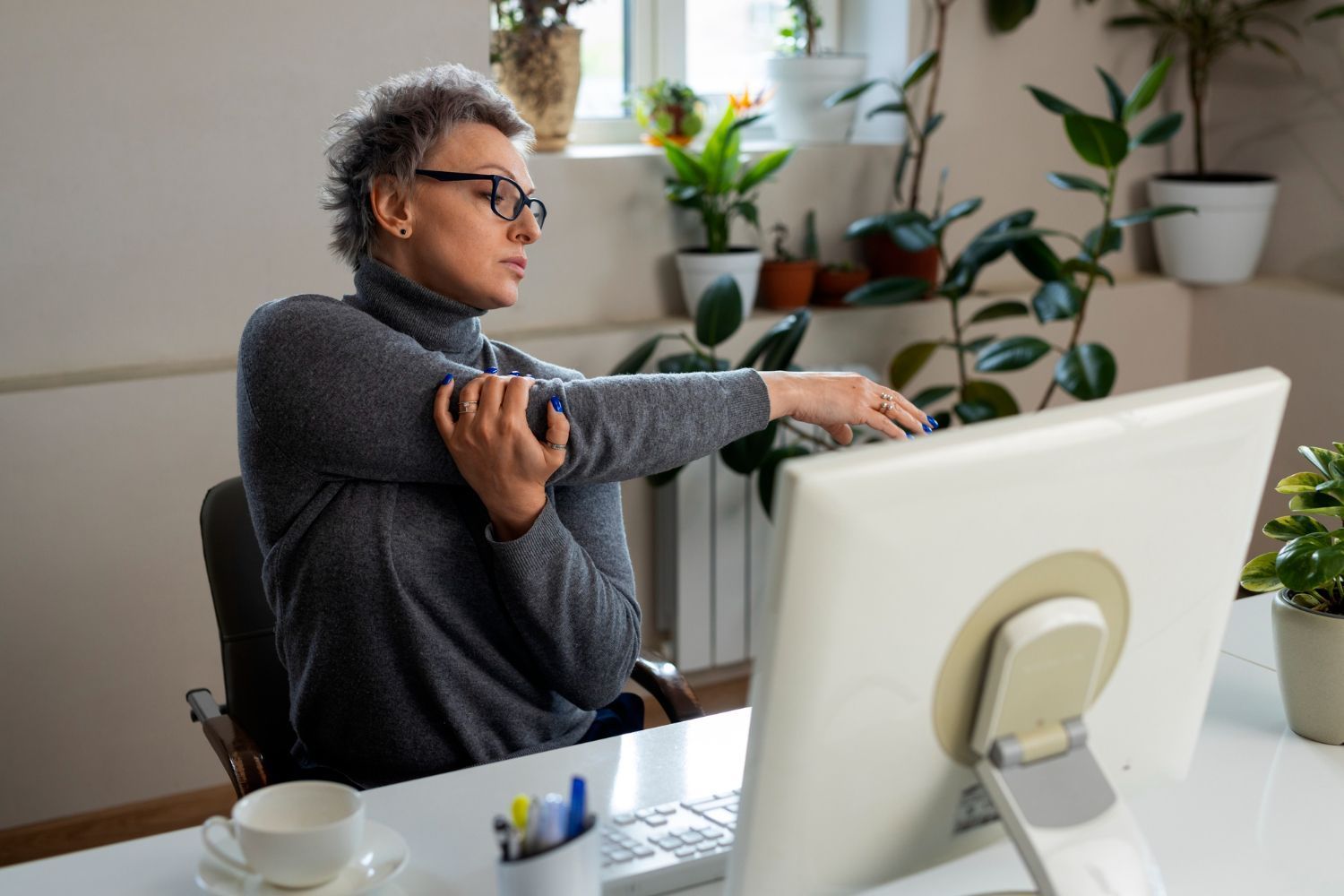 Woman in glasses stretches arm while seated at a computer, with plants in the background.