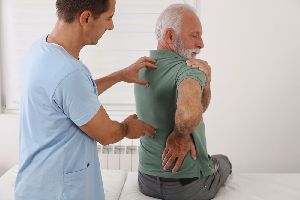 A physical therapist examines the back of a man sitting on a medical exam table.
