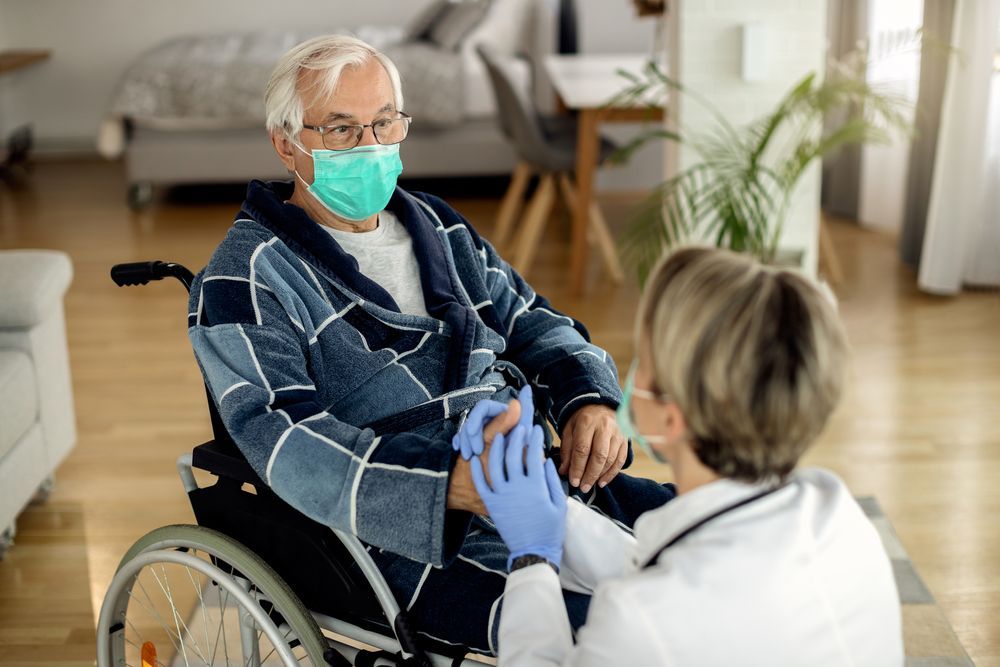 A healthcare professional in a white coat and blue gloves holds the hands of a person in a wheelchair wearing a face mask.