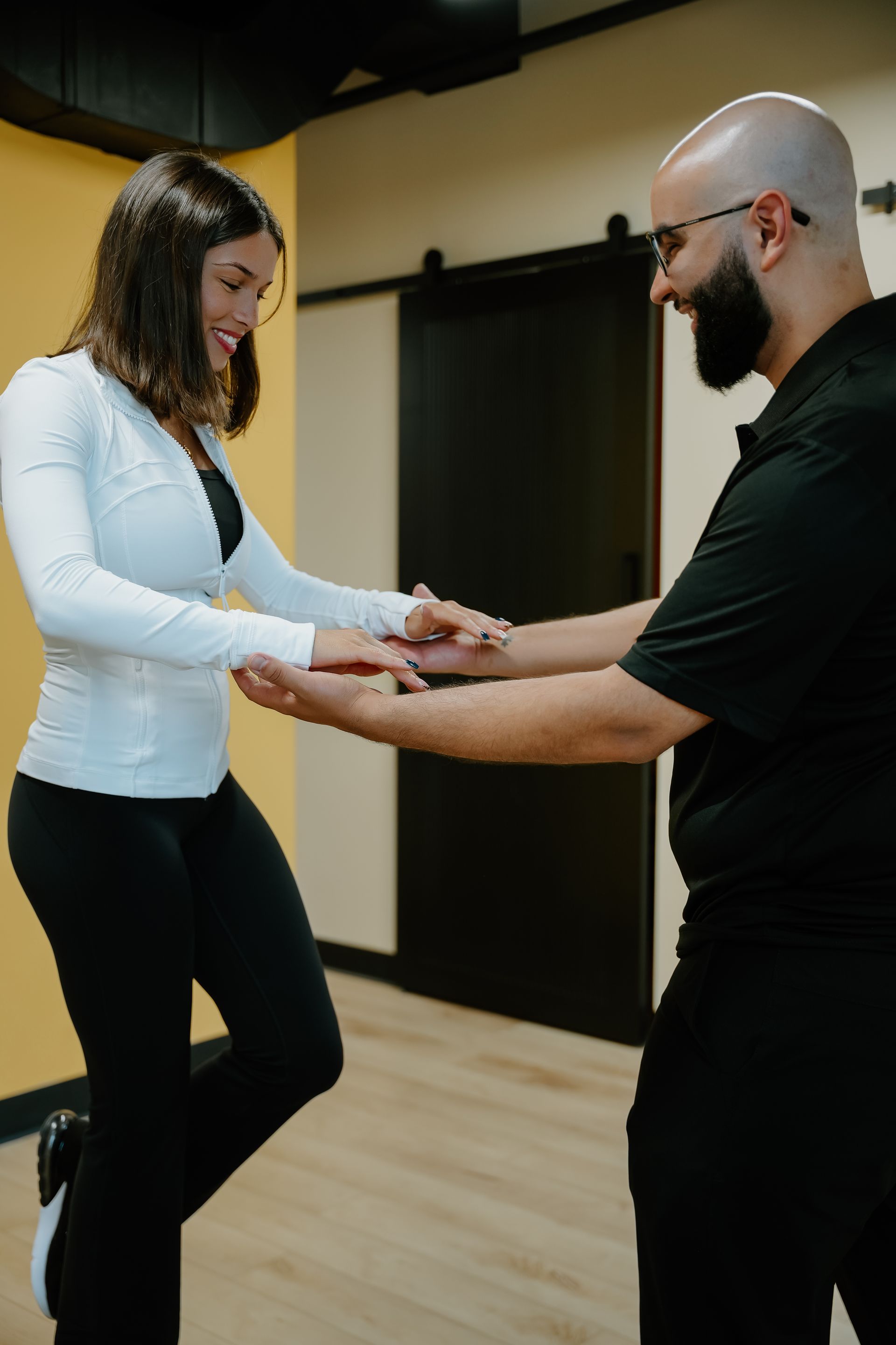 Woman balancing, assisted by a man, in a physical therapy setting. She smiles, holding his hands.
