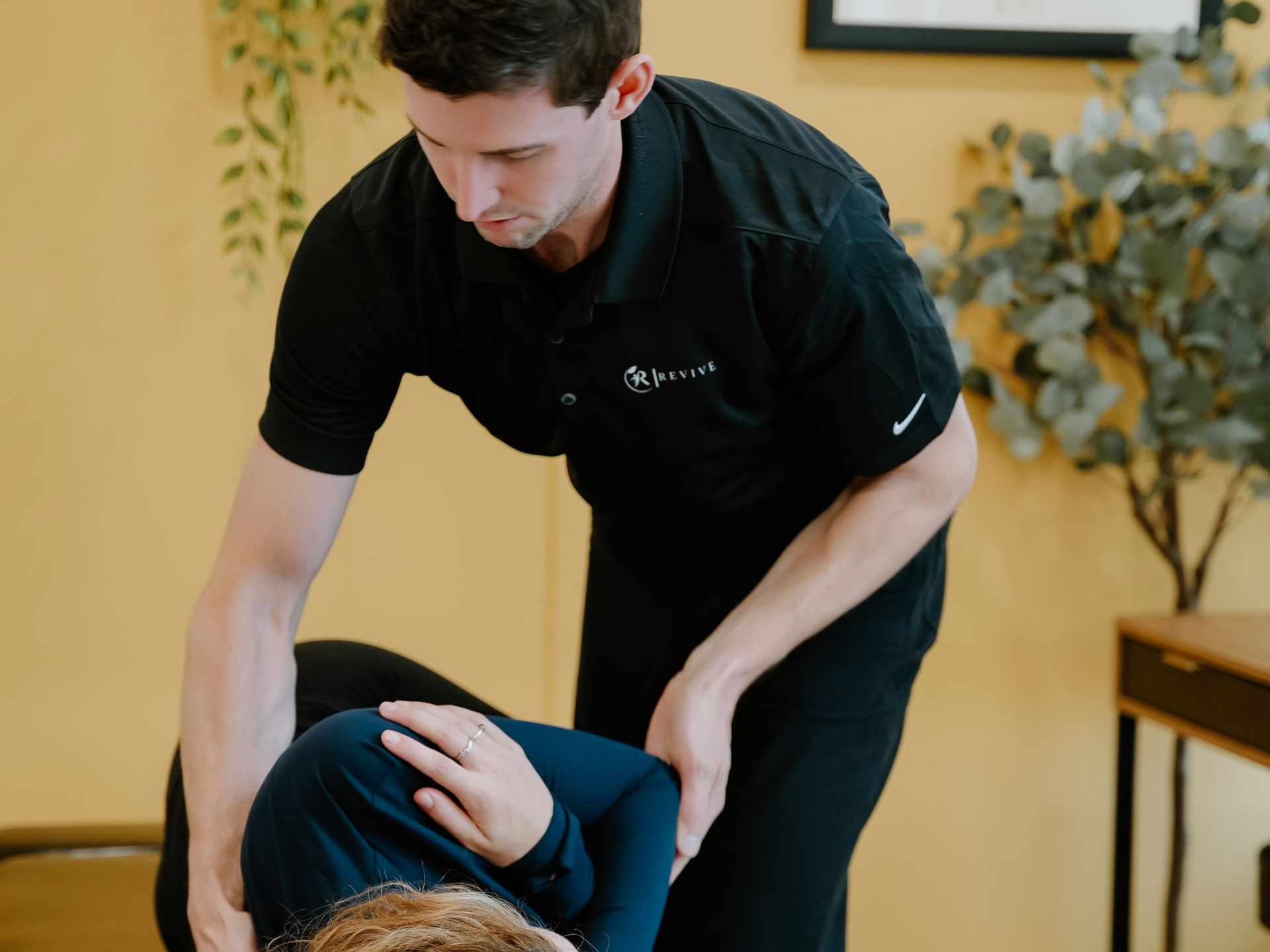 Chiropractor adjusting a patient's neck in a clinic. Both are dressed in dark clothing; yellow wall in the background.
