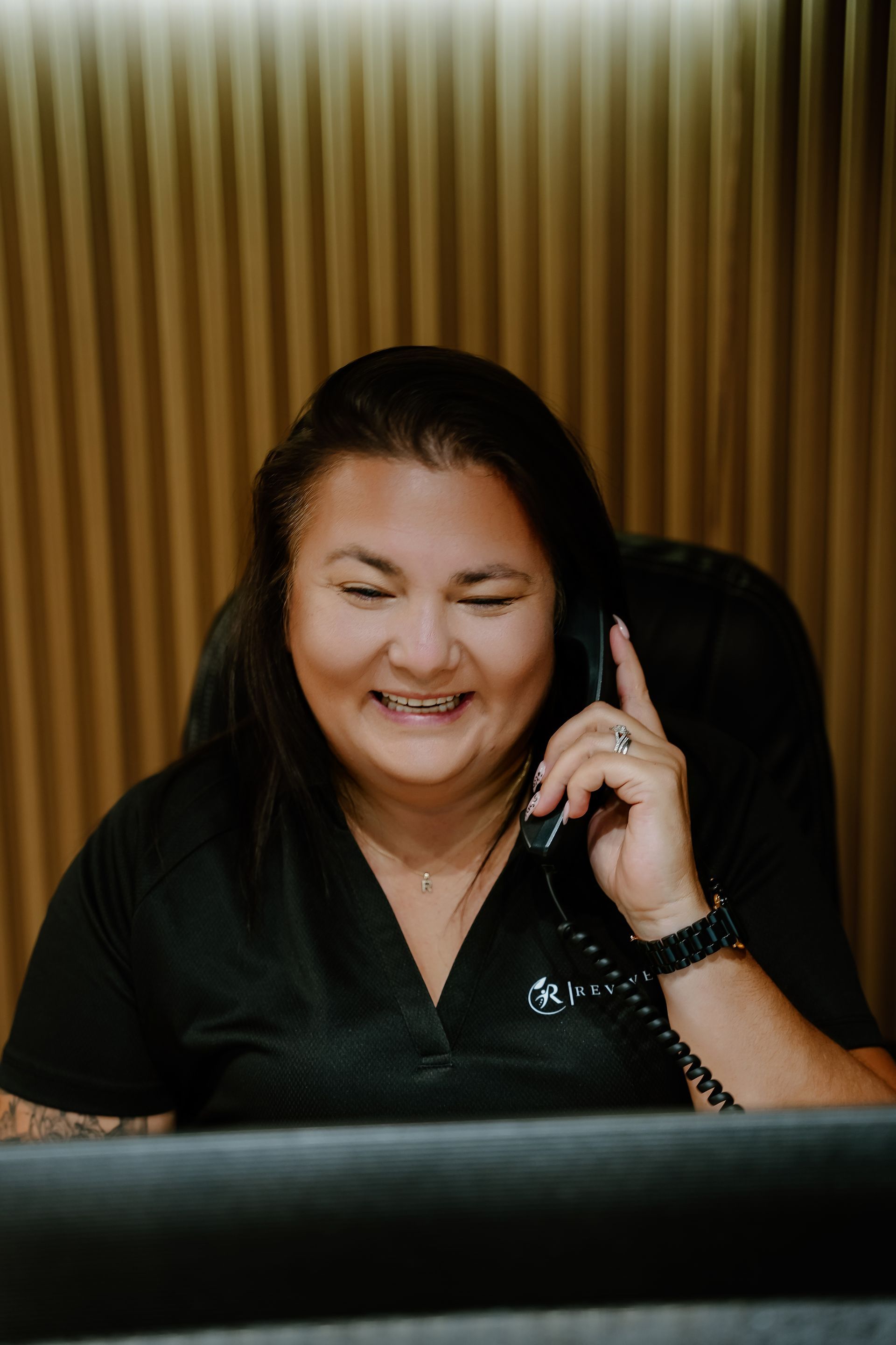Woman smiling, holding phone to ear, seated at desk in office, brown walls.