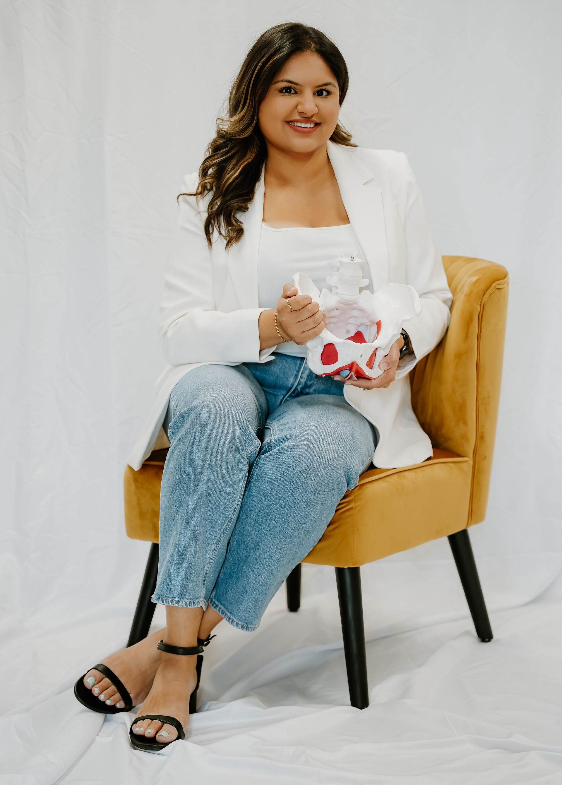 Woman in blazer and jeans sits holding a gift in an armchair, smiling, against a white backdrop.