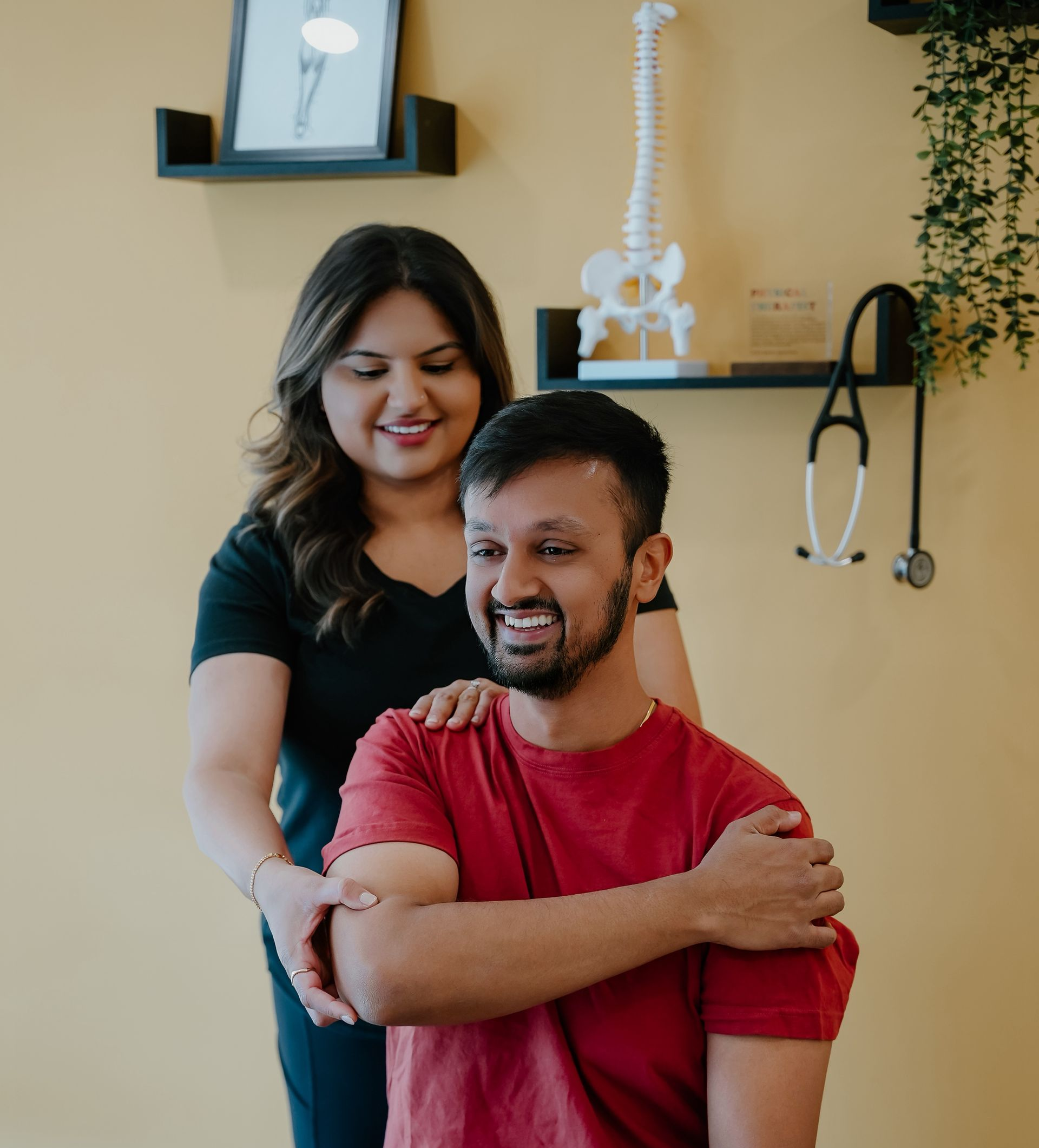 Woman adjusting man's arm in a chiropractic office. Both are smiling. Yellow wall, stethoscope, spine model visible.