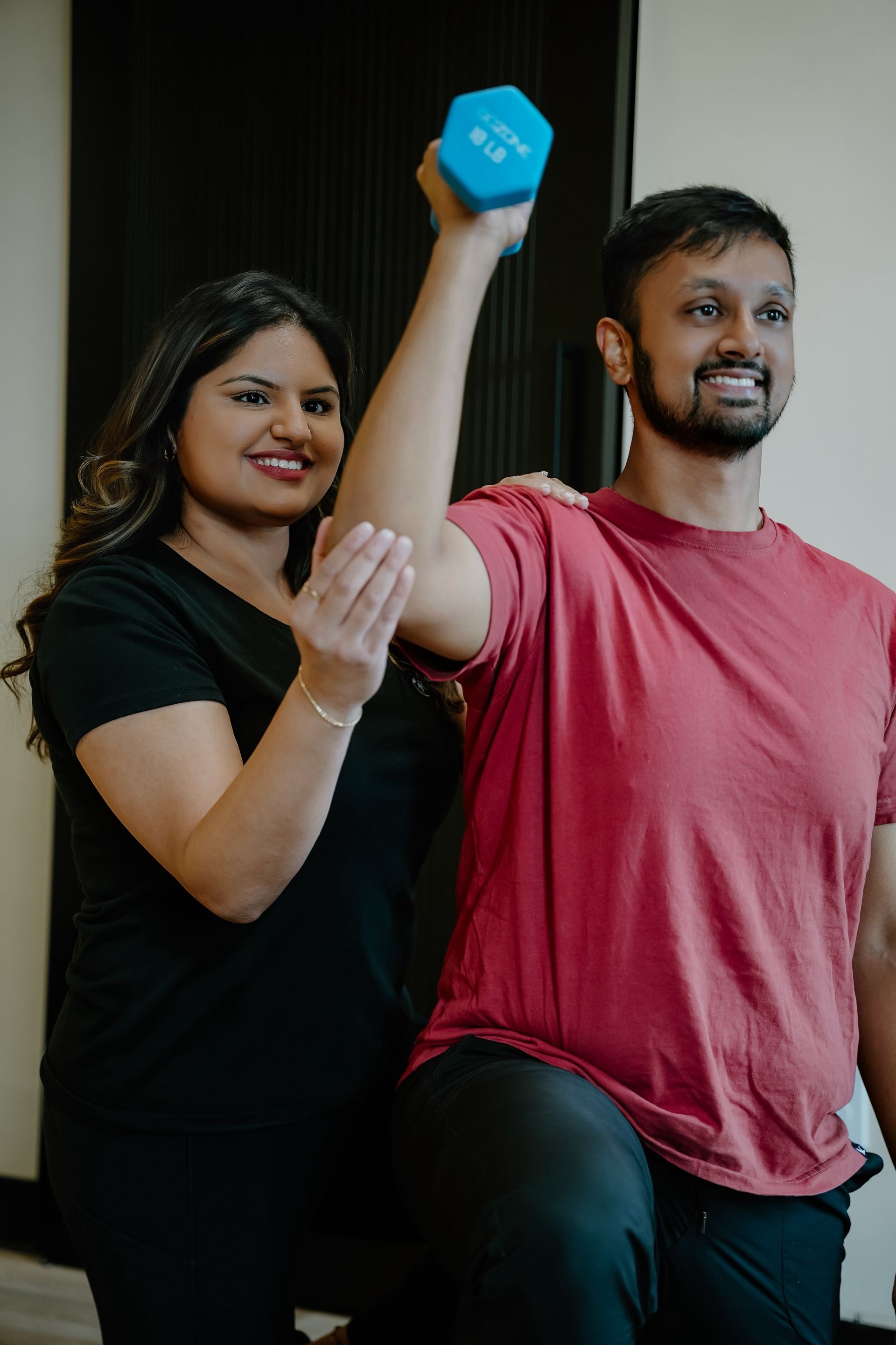 Woman assisting a person lifting a blue dumbbell. They're indoors. Person smiles. Woman has a hand on the arm.