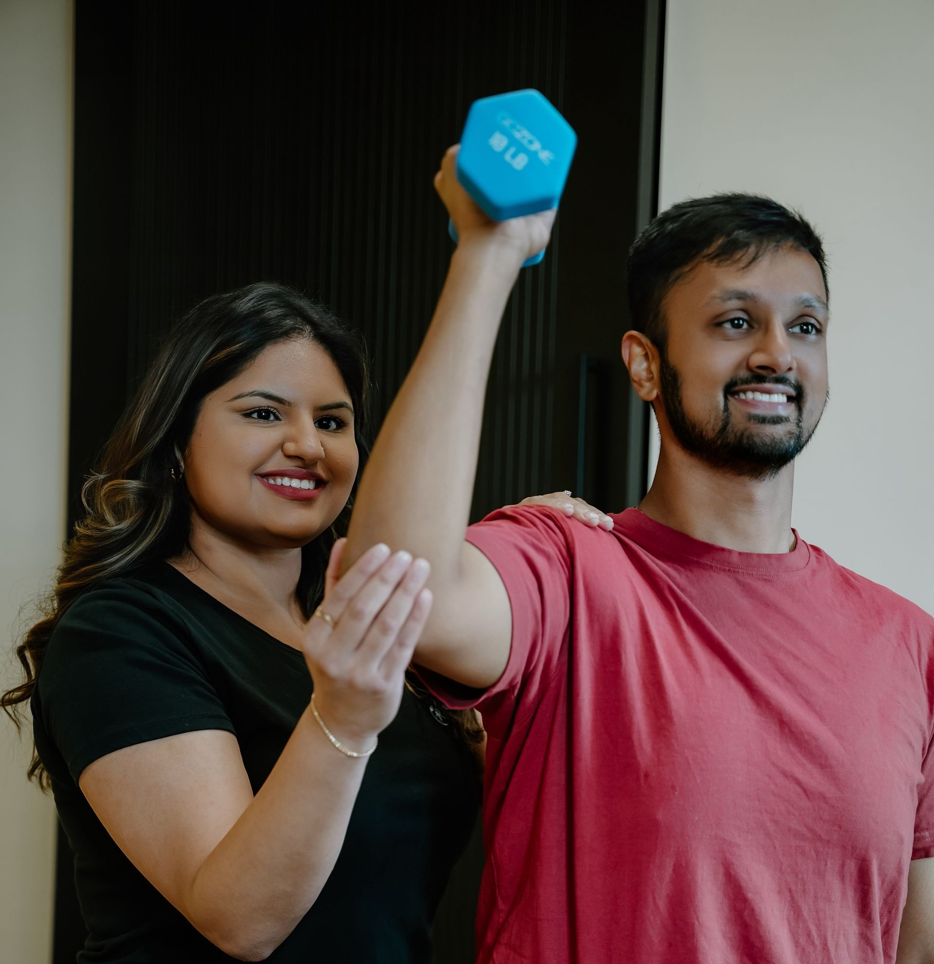 Woman assisting a person lifting a blue dumbbell. They're indoors. Person smiles. Woman has a hand on the arm.