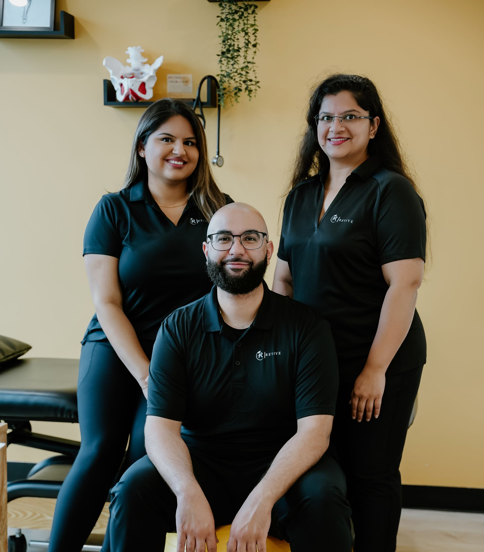 Three healthcare professionals pose for a photo in a yellow-walled office. The man in front is bald and bearded; the women smile on either side.