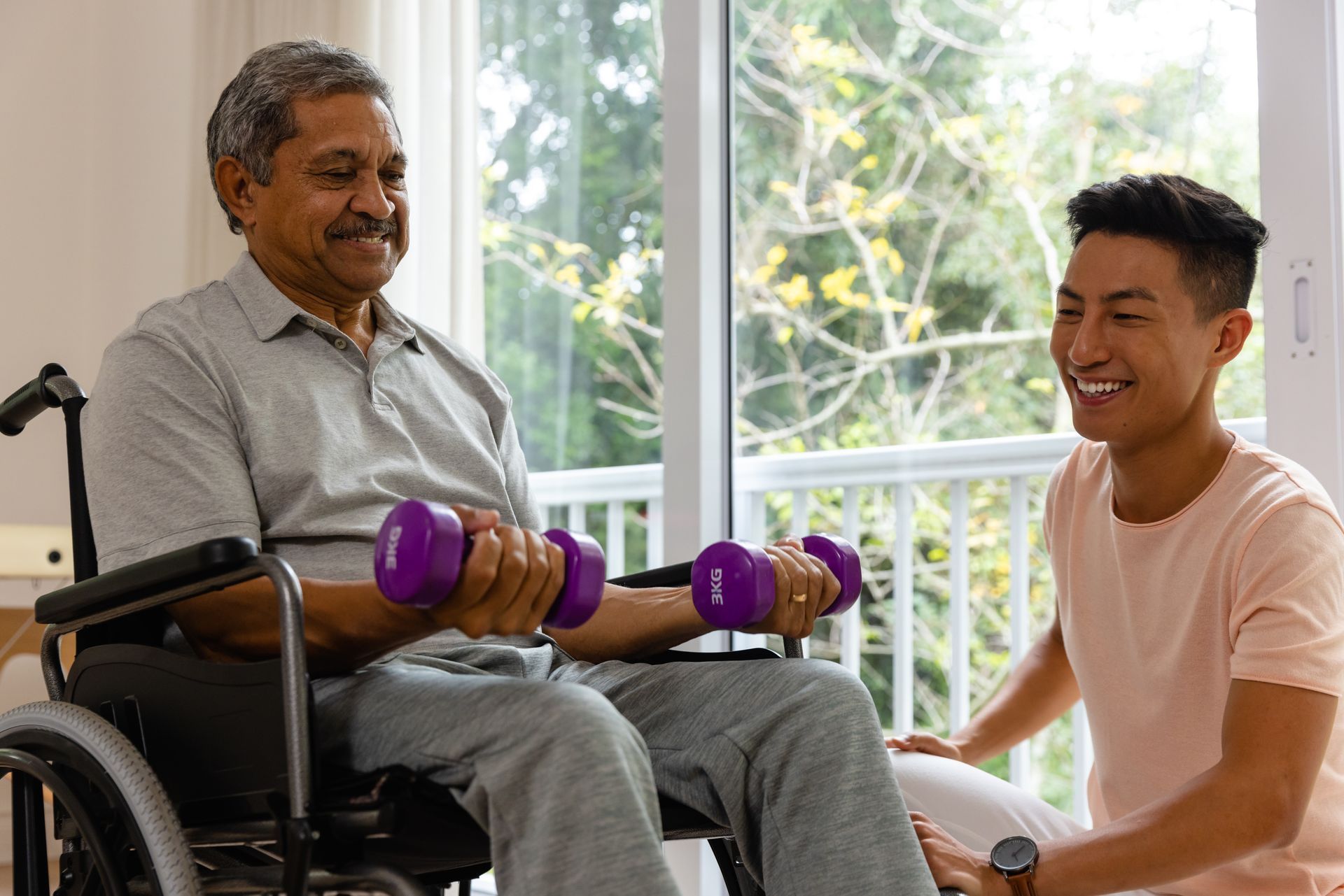 Man in wheelchair lifts dumbbells with assistance from a smiling person near a window.