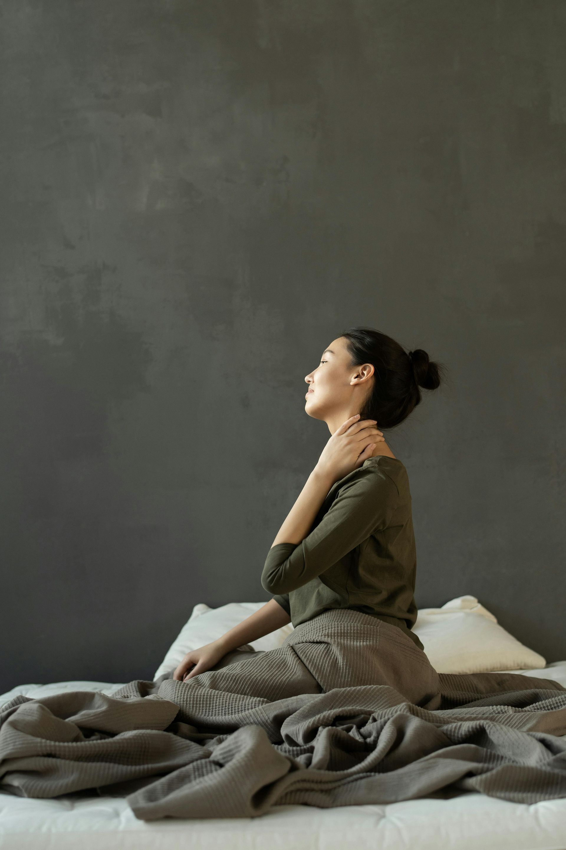 Woman sitting on a bed, hand on neck, looking up. Dark gray wall backdrop, gray blanket, pillows.