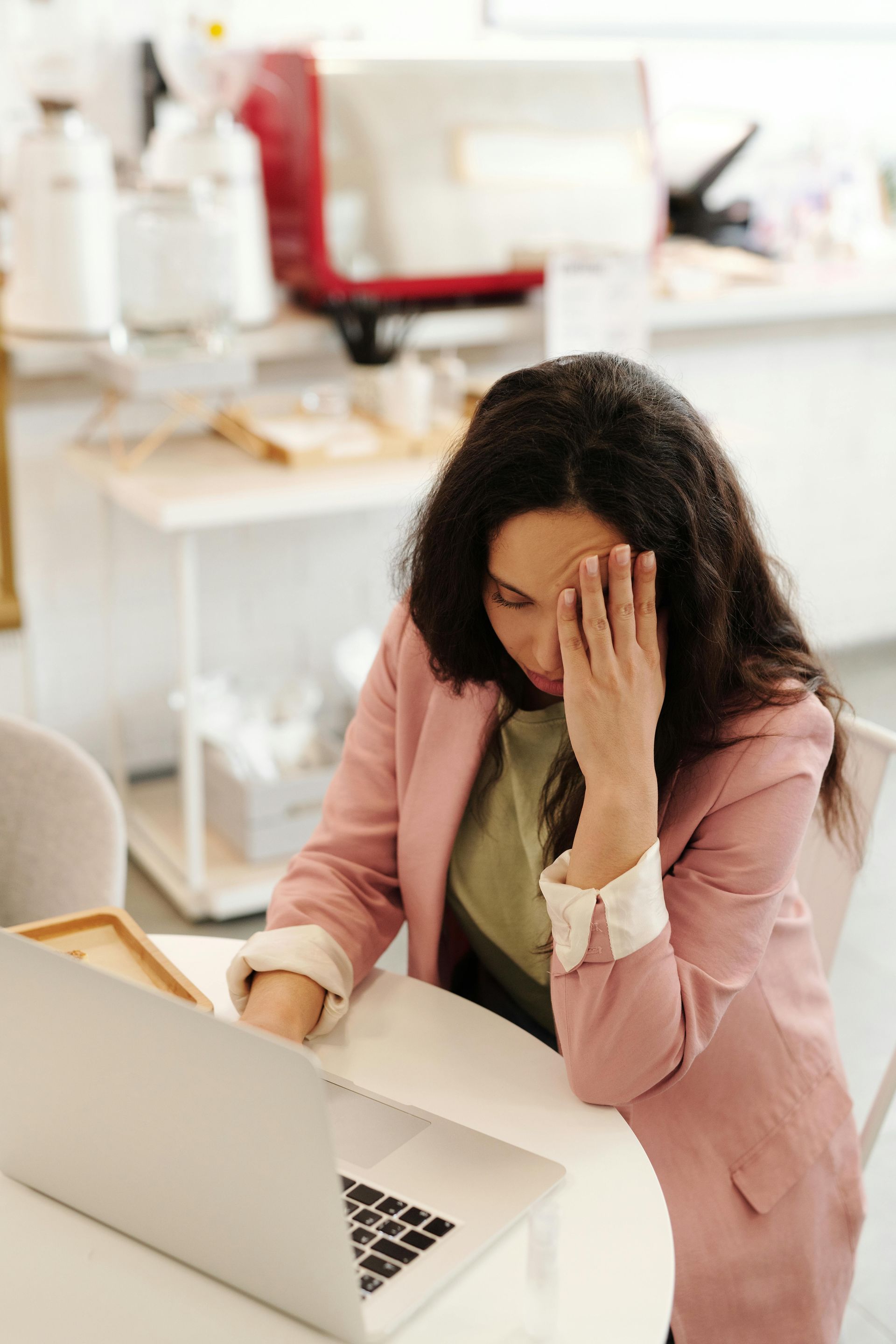 Woman with hand on forehead, looking at laptop; pink blazer in a light-filled room.