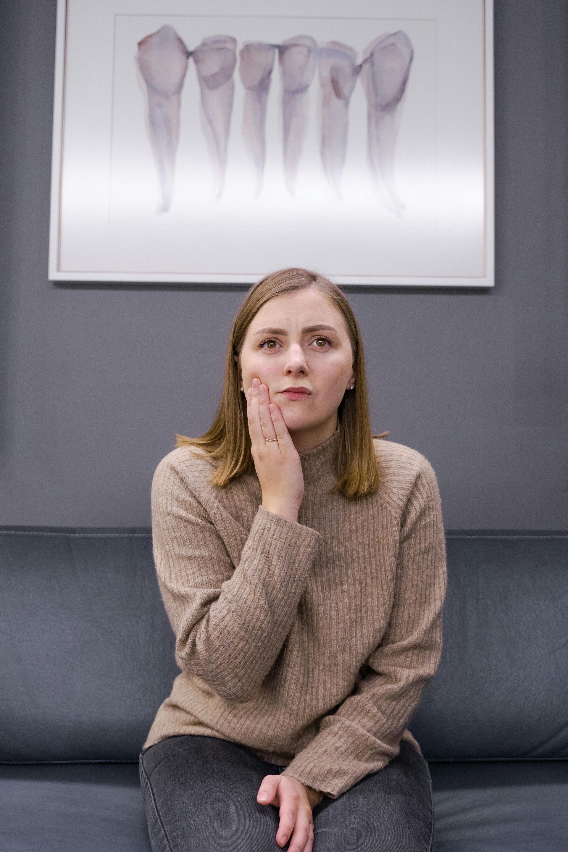 Woman holding cheek, possibly in pain, seated in a dental office, with teeth art on the wall.