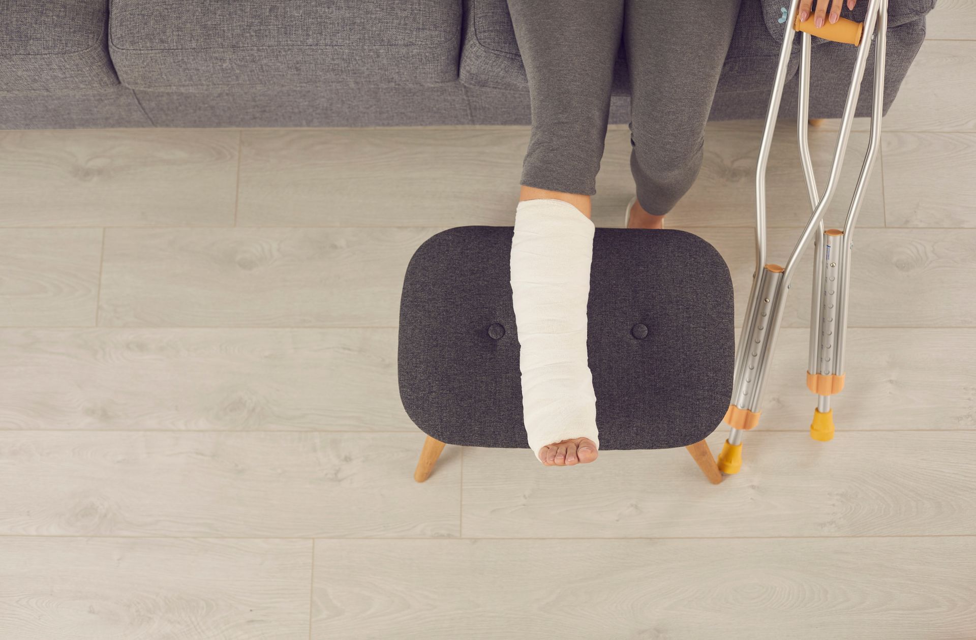 Person with a leg in a cast rests foot on a stool, crutches nearby, sitting on a sofa.