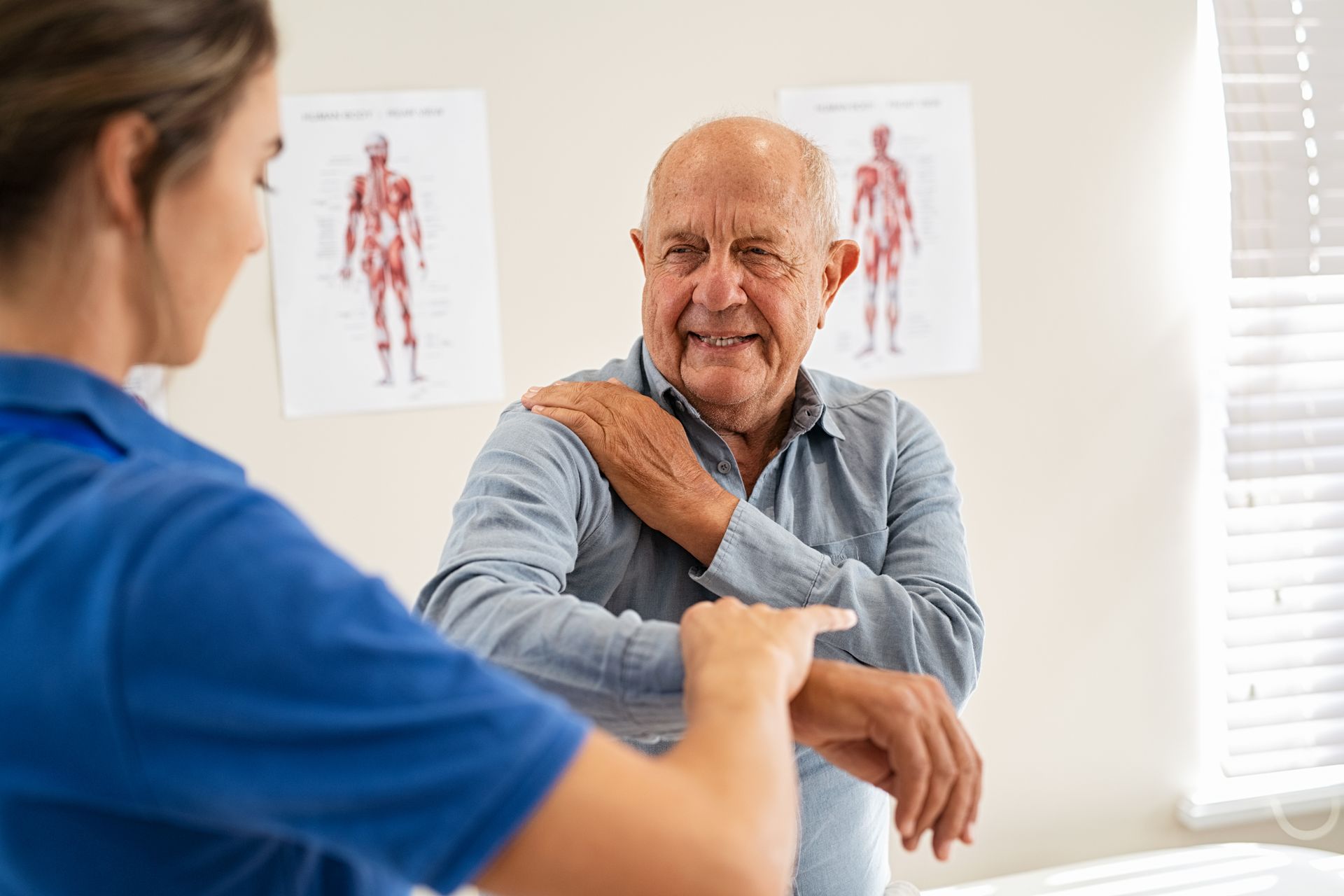 A person in a blue uniform examining the shoulder of an individual in a clinical setting with anatomical charts.