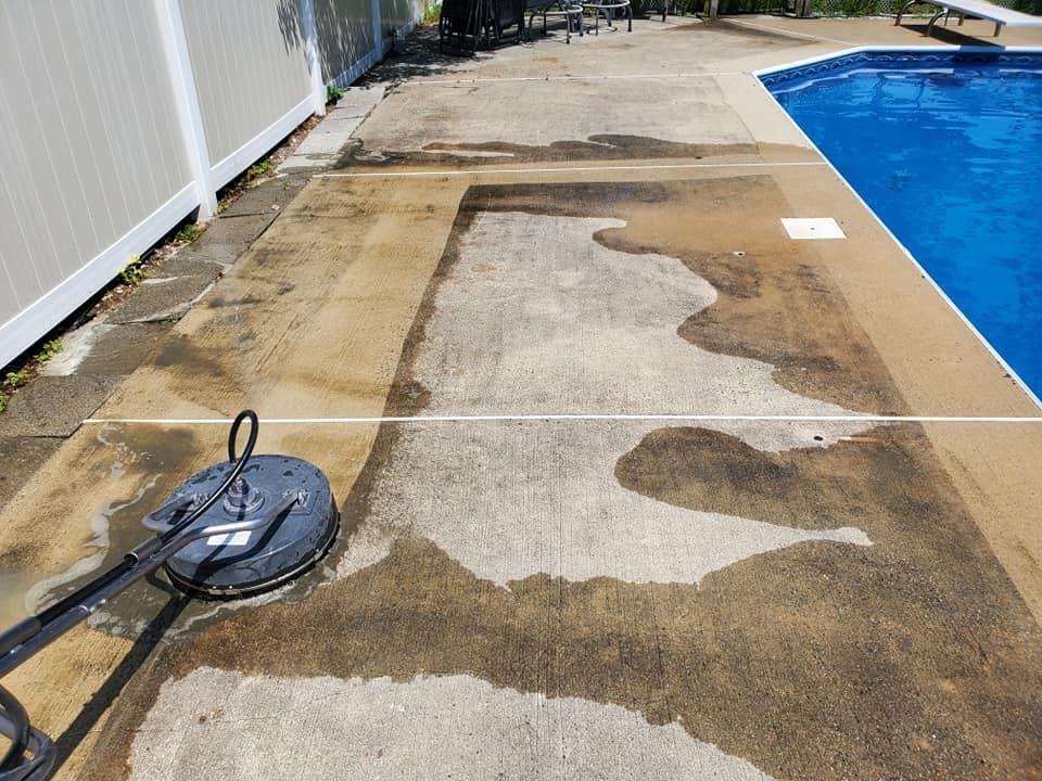 A concrete patio next to a swimming pool is being cleaned with a pressure washer.