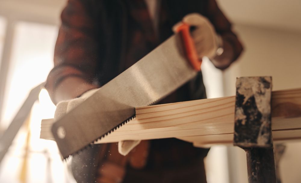 Person sawing wood with a hand saw, wearing a plaid shirt in a workshop.