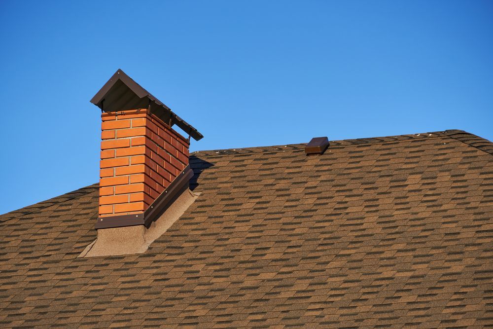 Brick chimney on a brown shingled roof against a clear blue sky.