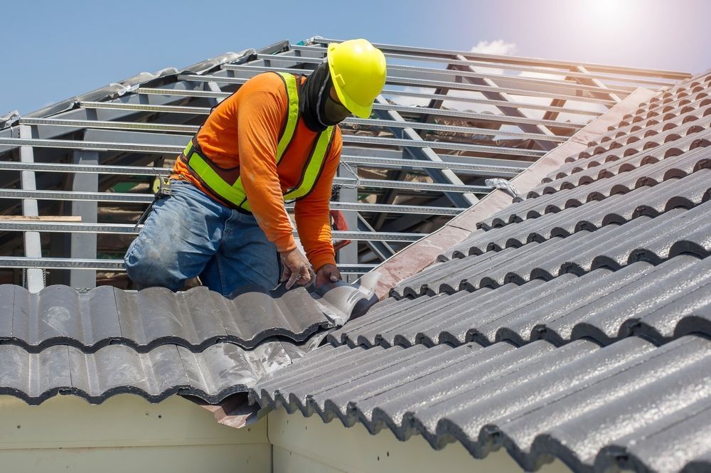 Roofer in orange shirt and yellow helmet working on a tiled roof under a blue sky.