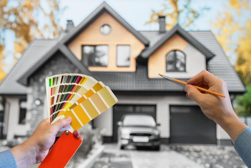 Hands holding paint swatches and brush, modifying a grayscale house exterior with color.