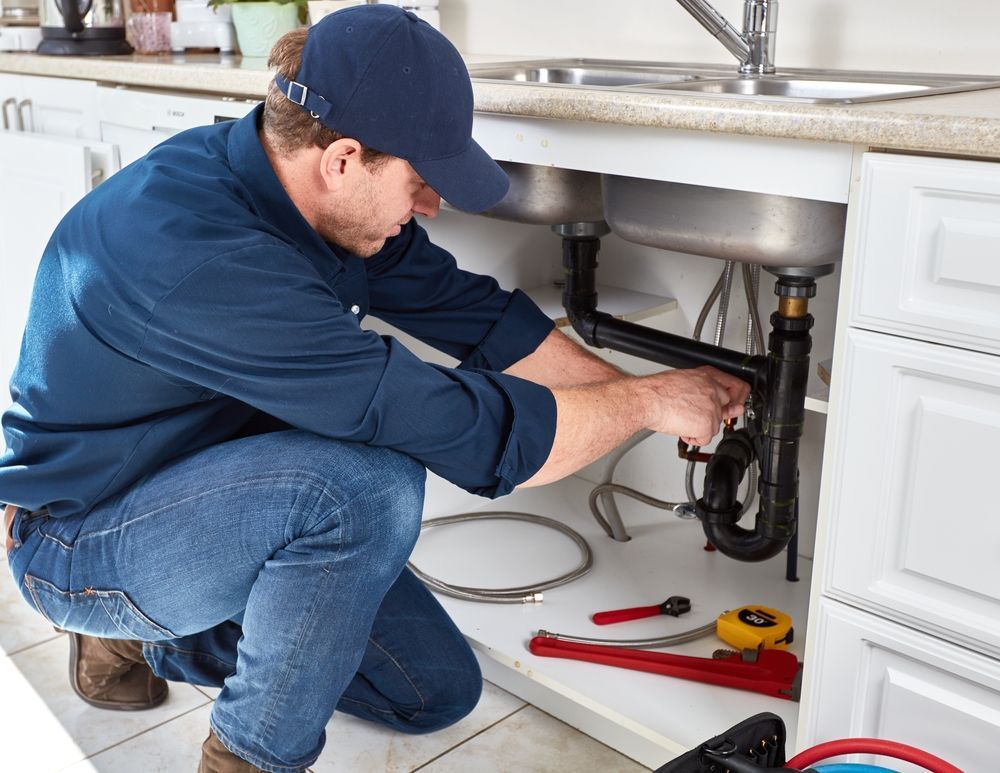 Plumber in blue hat and shirt working under a kitchen sink with tools; indoor setting.