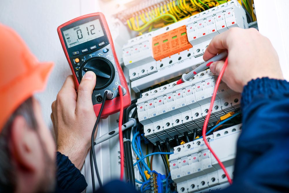 Electrician testing an electrical panel with a multimeter, wearing an orange helmet and blue jacket.