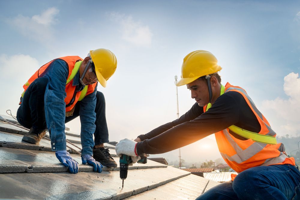 Two roofers in hard hats and vests working on a rooftop, one using a drill.