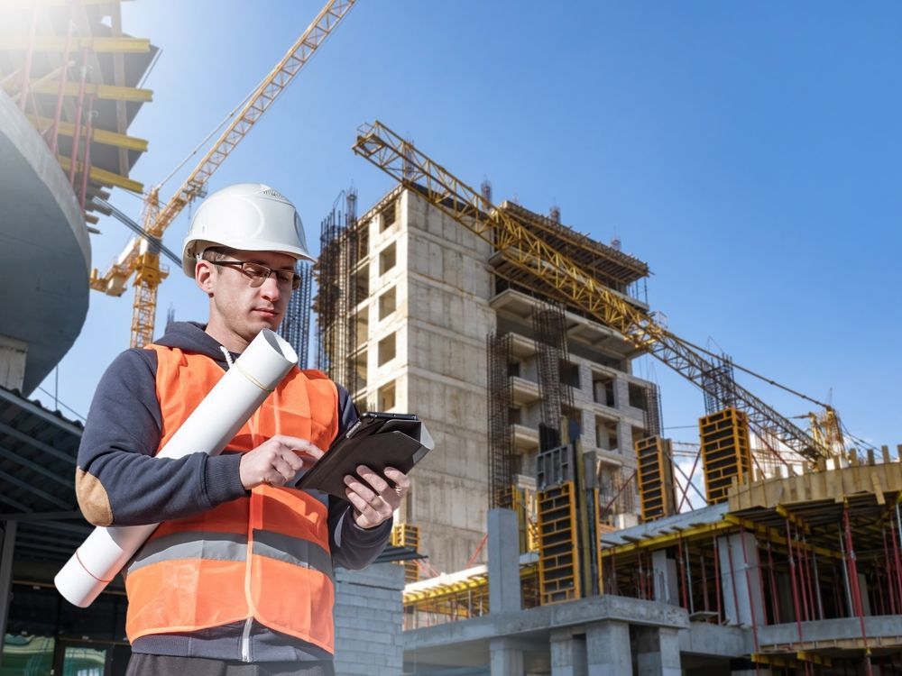 Construction worker on site, orange vest, hard hat, using tablet, holding blueprints, building in background.