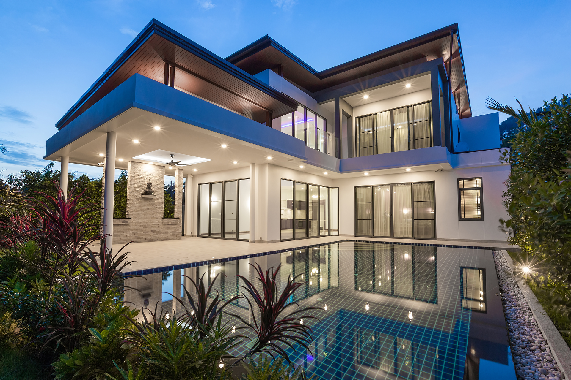 Modern multi-story house with pool, glass windows, and outdoor lighting, reflecting in the water at dusk.