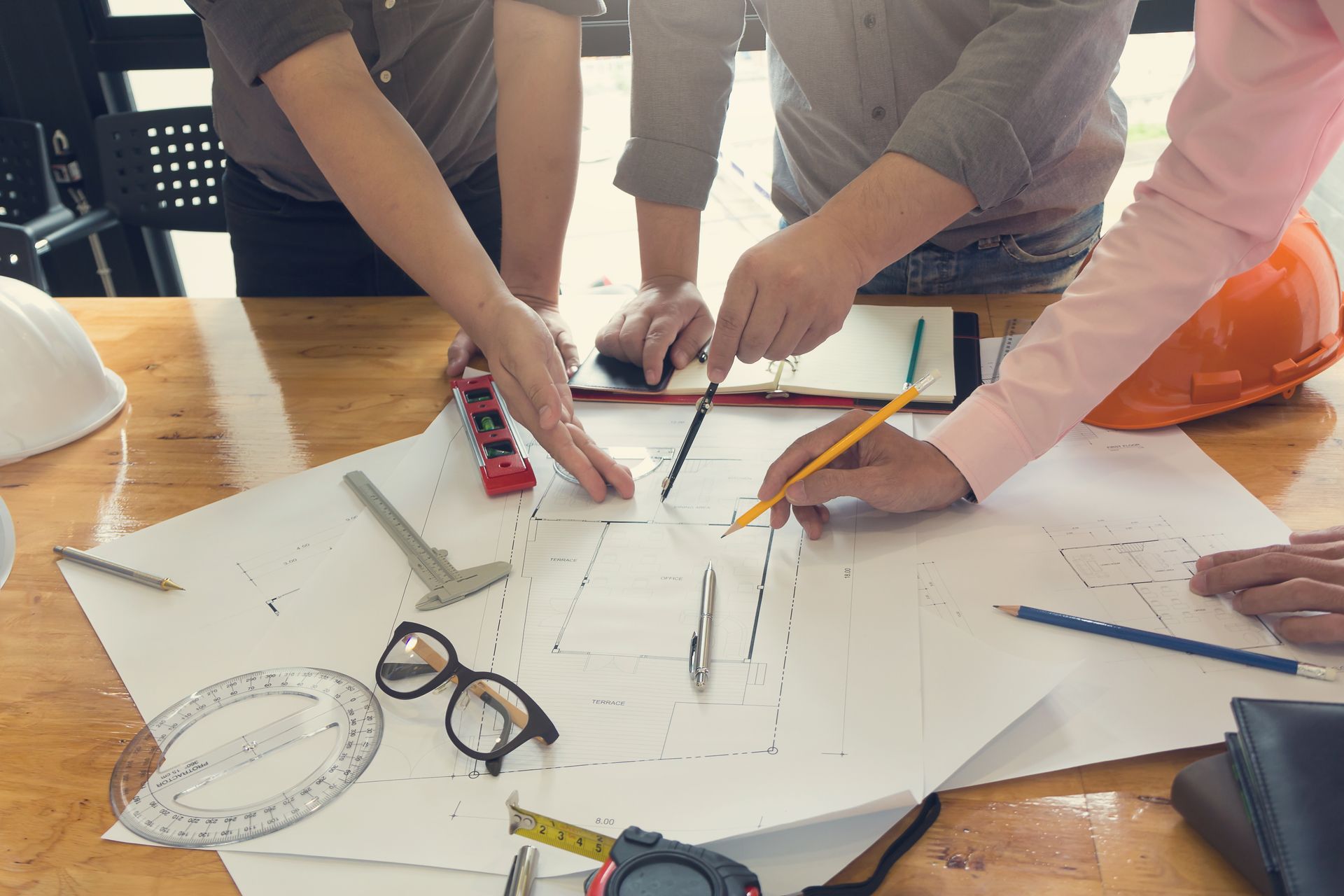 Persone che collaborano attorno a progetti su un tavolo di legno. Sono presenti attrezzi e un casco protettivo.