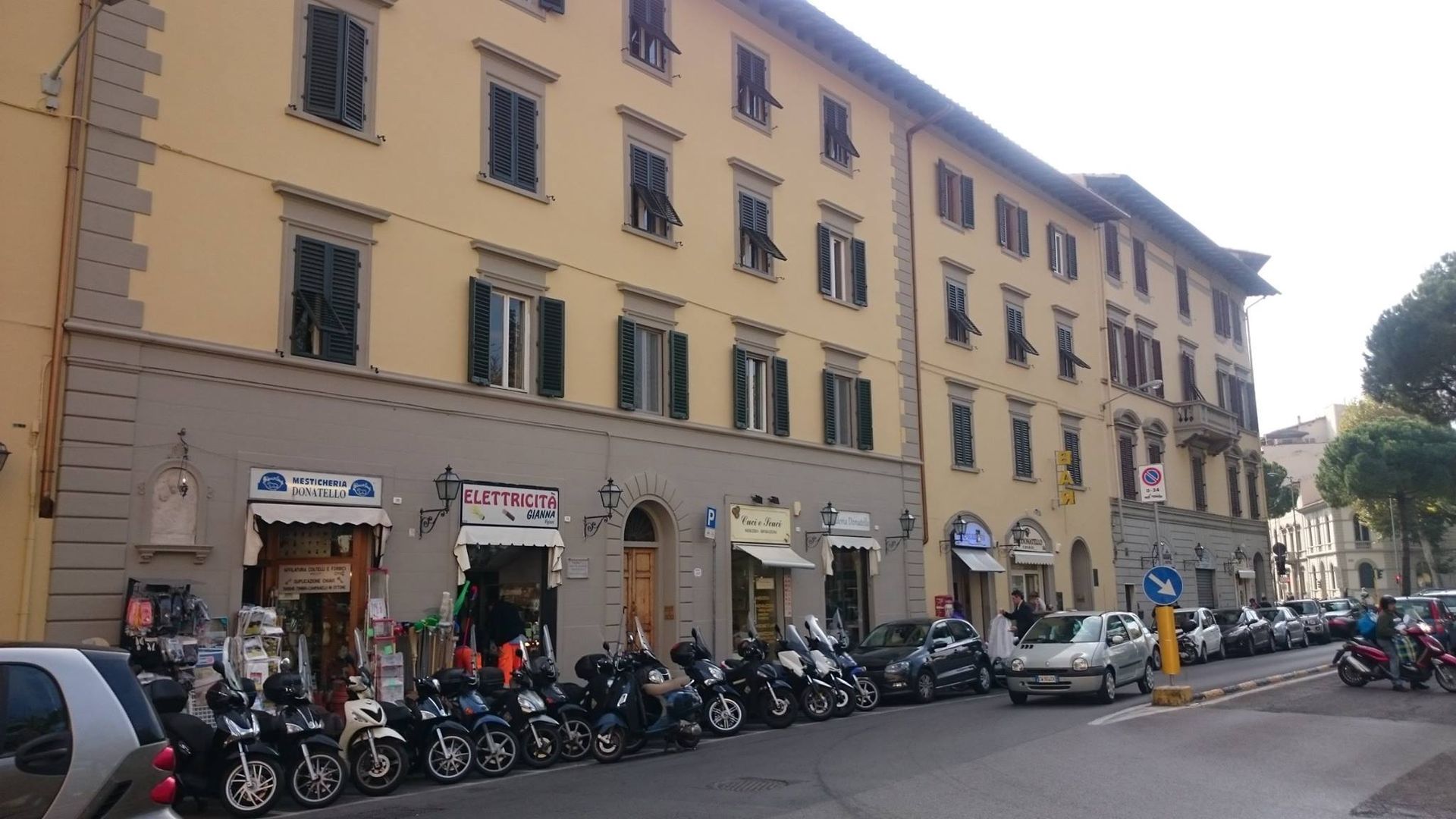 Una fila di negozi e motociclette parcheggiate costeggiano una strada di fronte a un edificio giallo chiaro.
