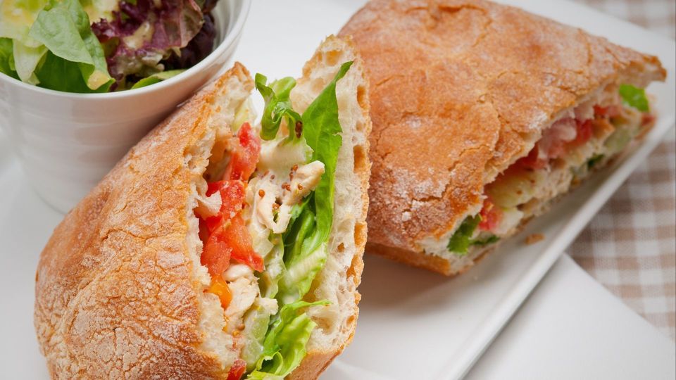 Ciabatta sandwich, cut in half, filled with lettuce, tomato, and what appears to be chicken. Served with a side salad.