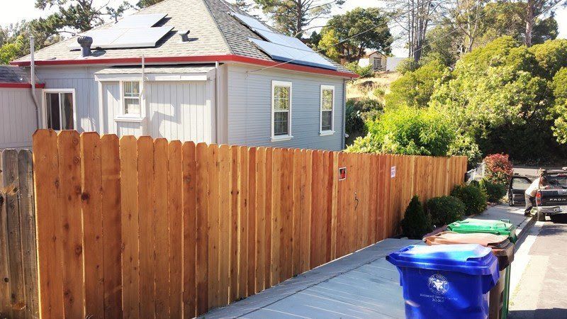 Wooden fence bordering a light blue house with solar panels on its roof, next to a street with recycling bins.