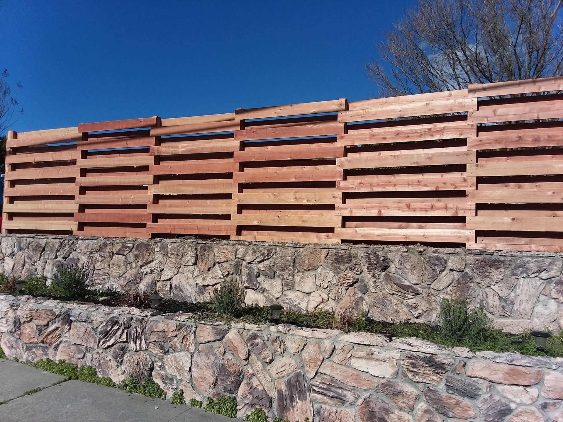 Wooden slat fence atop a stone retaining wall against a blue sky.
