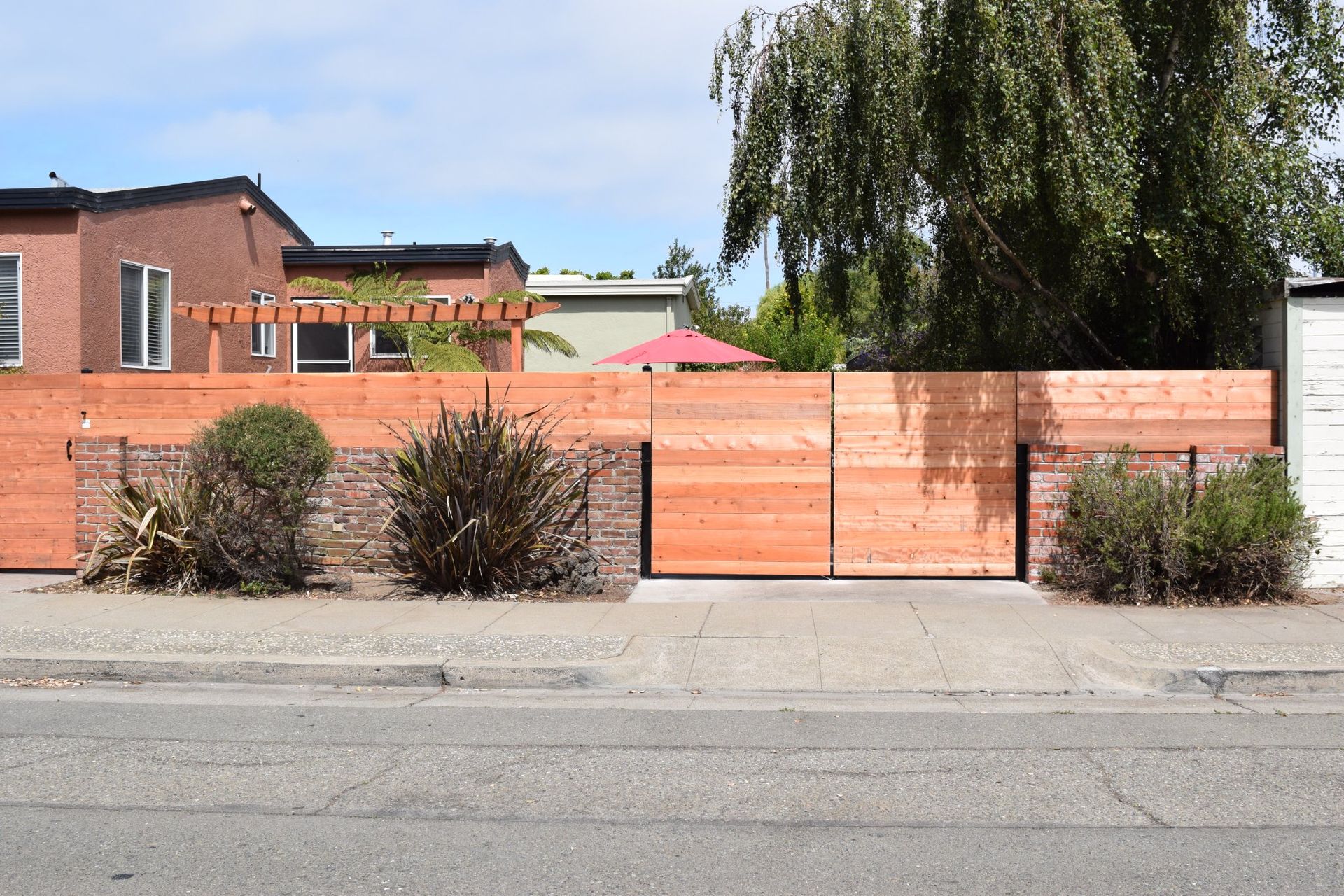 Wooden fence with gate, greenery, and houses along a sidewalk.
