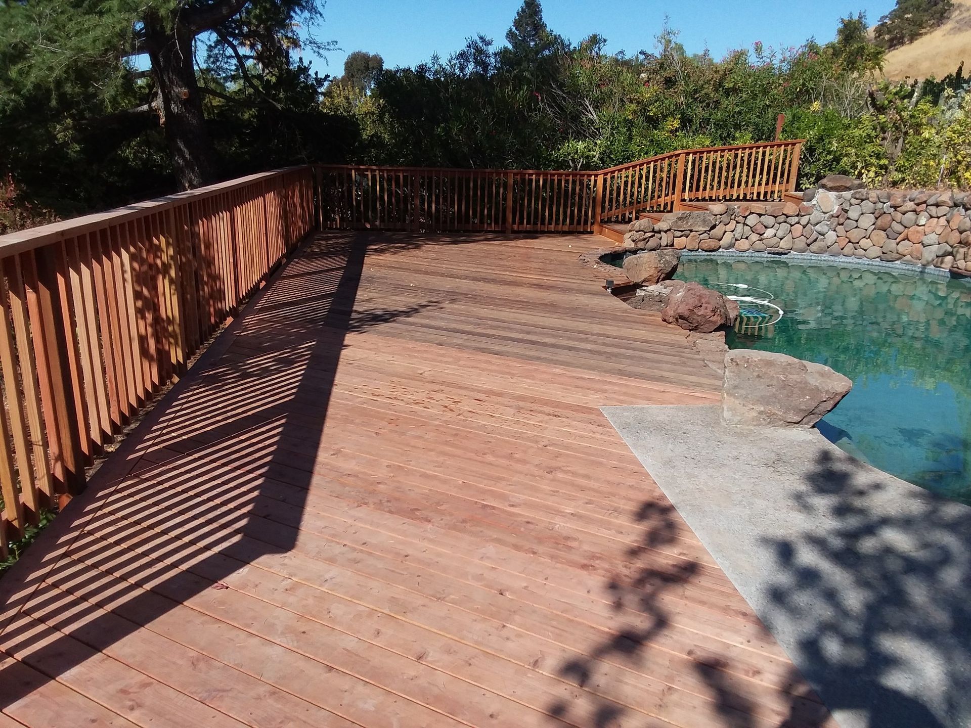 Wooden deck with railings overlooks a swimming pool on a sunny day.
