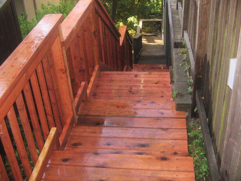 Wooden outdoor staircase with railings, stained reddish-brown. Descending into a partially wooded area.