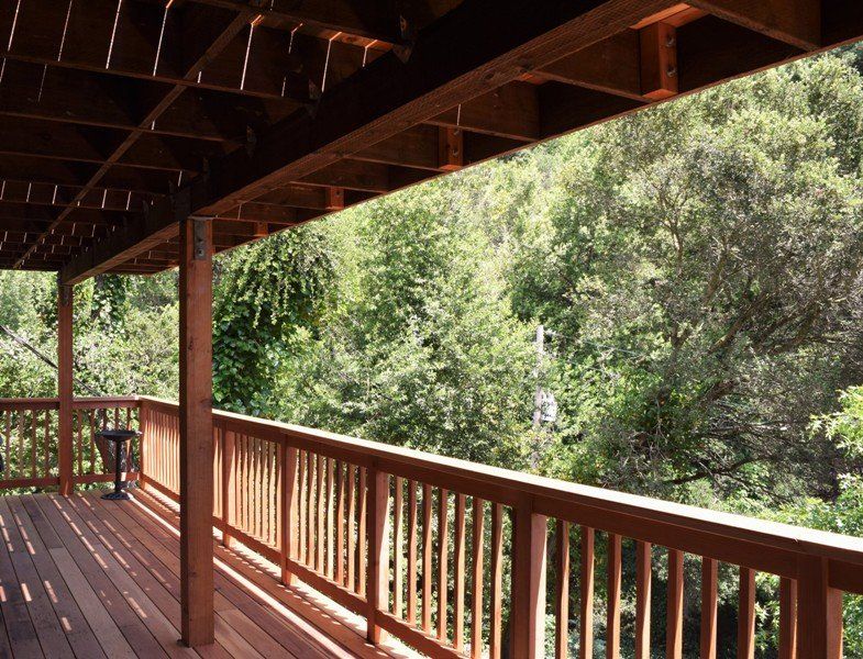 Wooden deck with railing overlooking a lush green forest, brown wood beams overhead.