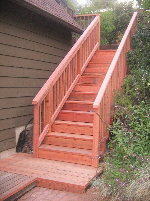 Wooden outdoor staircase with railings, leading up to a house. Green foliage and wood siding are visible.