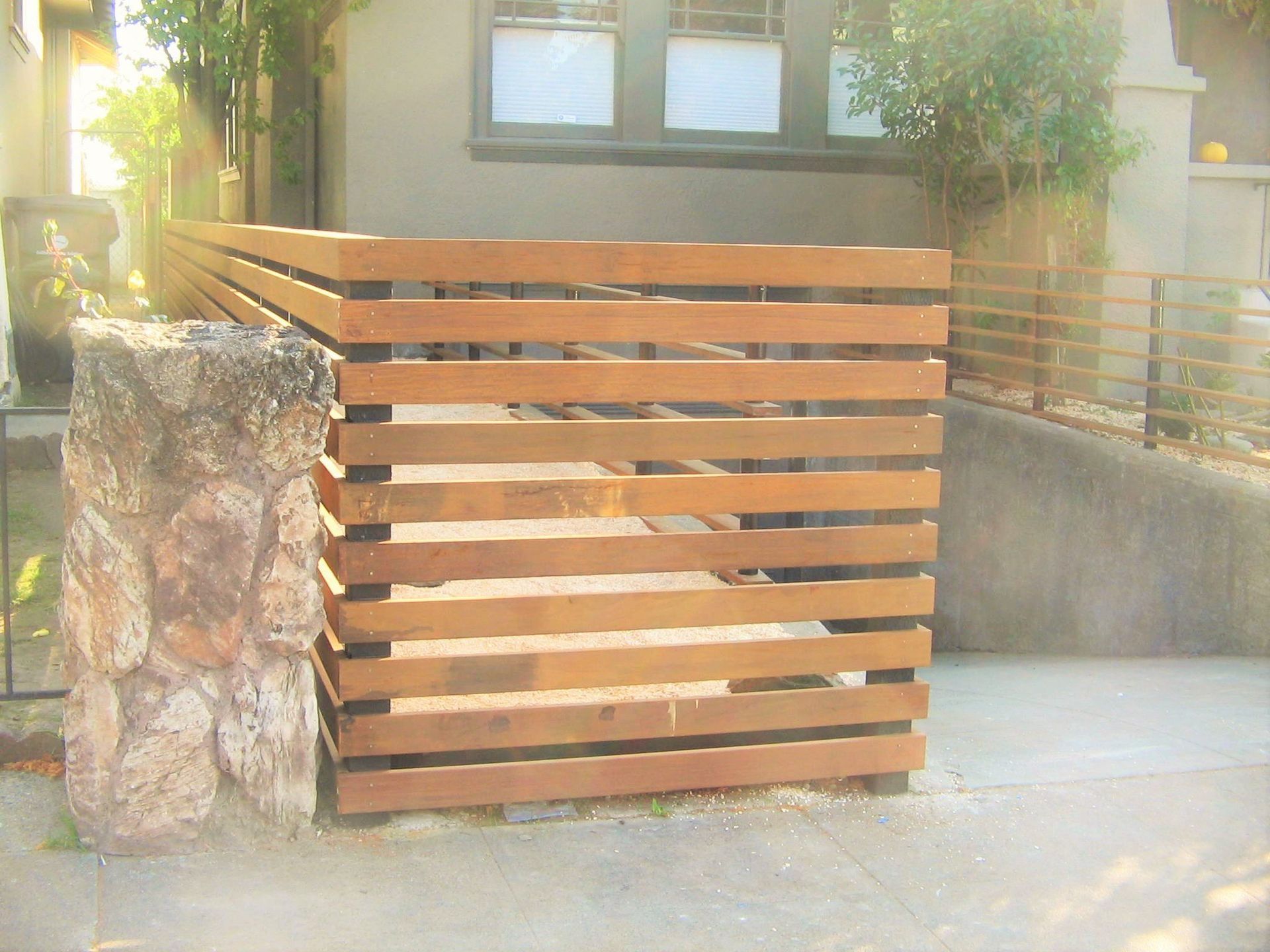 Wooden horizontal slat fence in front of a building next to a stone pillar, on a sidewalk.