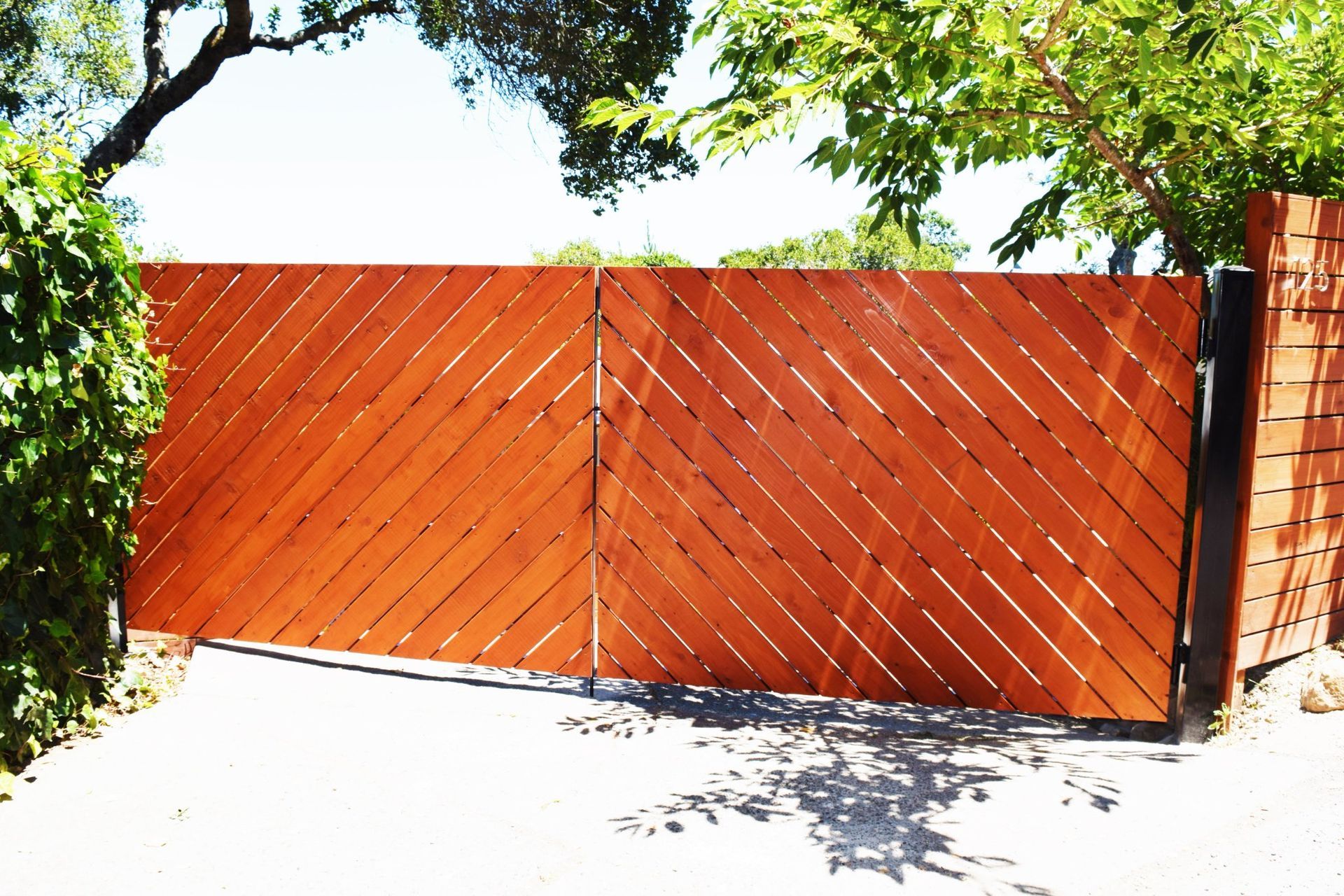 Wooden gate with diagonal slats, leading into a driveway, surrounded by greenery.