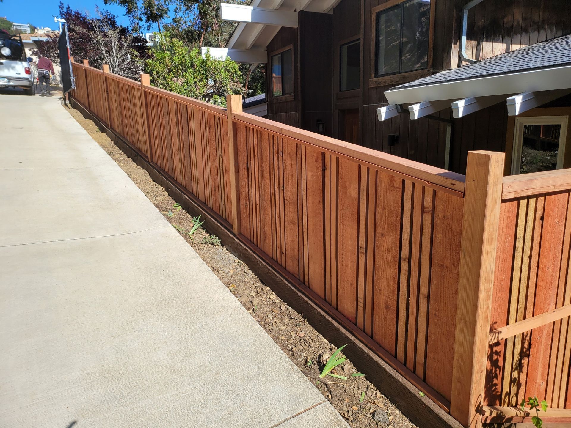 Wooden fence along a sloping concrete driveway next to a brown house.