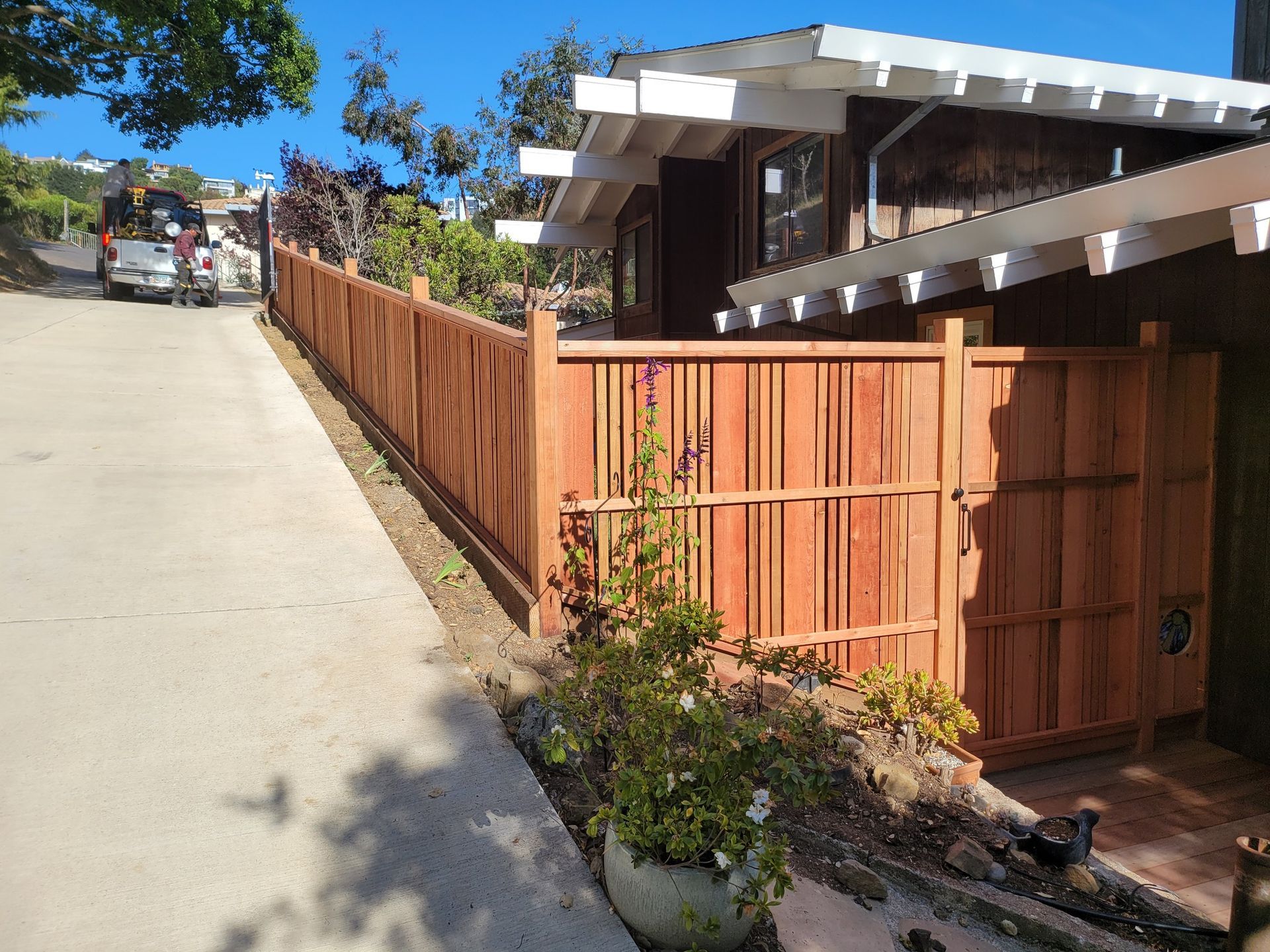 Wooden fence alongside a sloping driveway next to a brown house with a white roof.