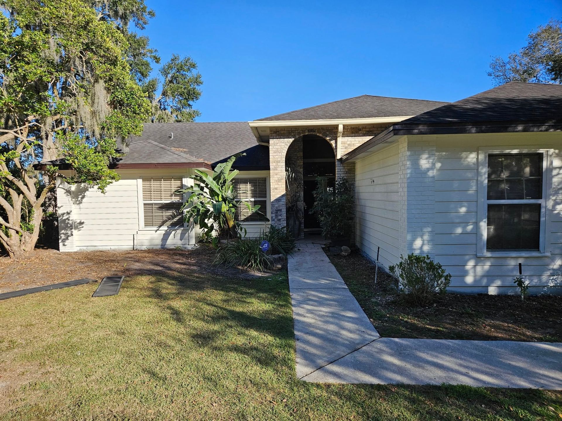 Ranch-style house with beige siding, a dark roof, and a walkway leading to the front door. Green lawn, blue sky.