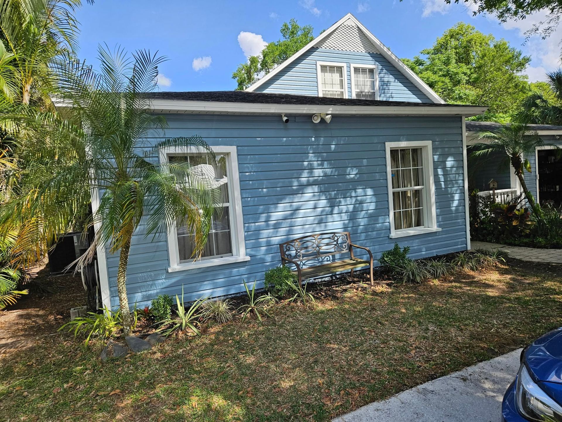 Blue house with white-trimmed windows, bench, and greenery. Sunny day.