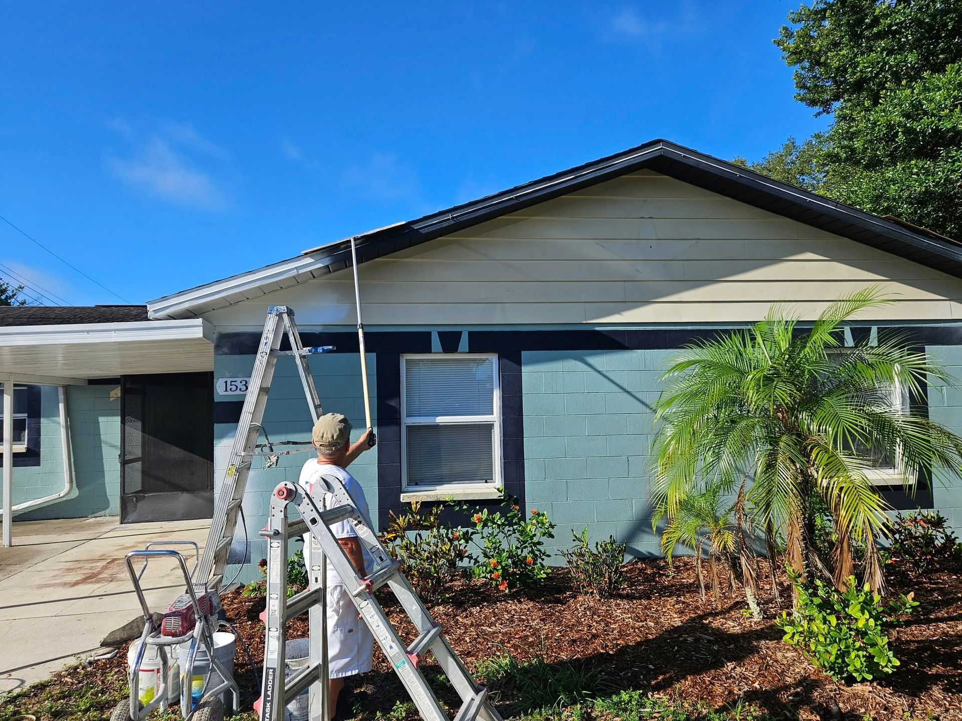 Person on ladder painting a teal and tan house exterior under a blue sky.